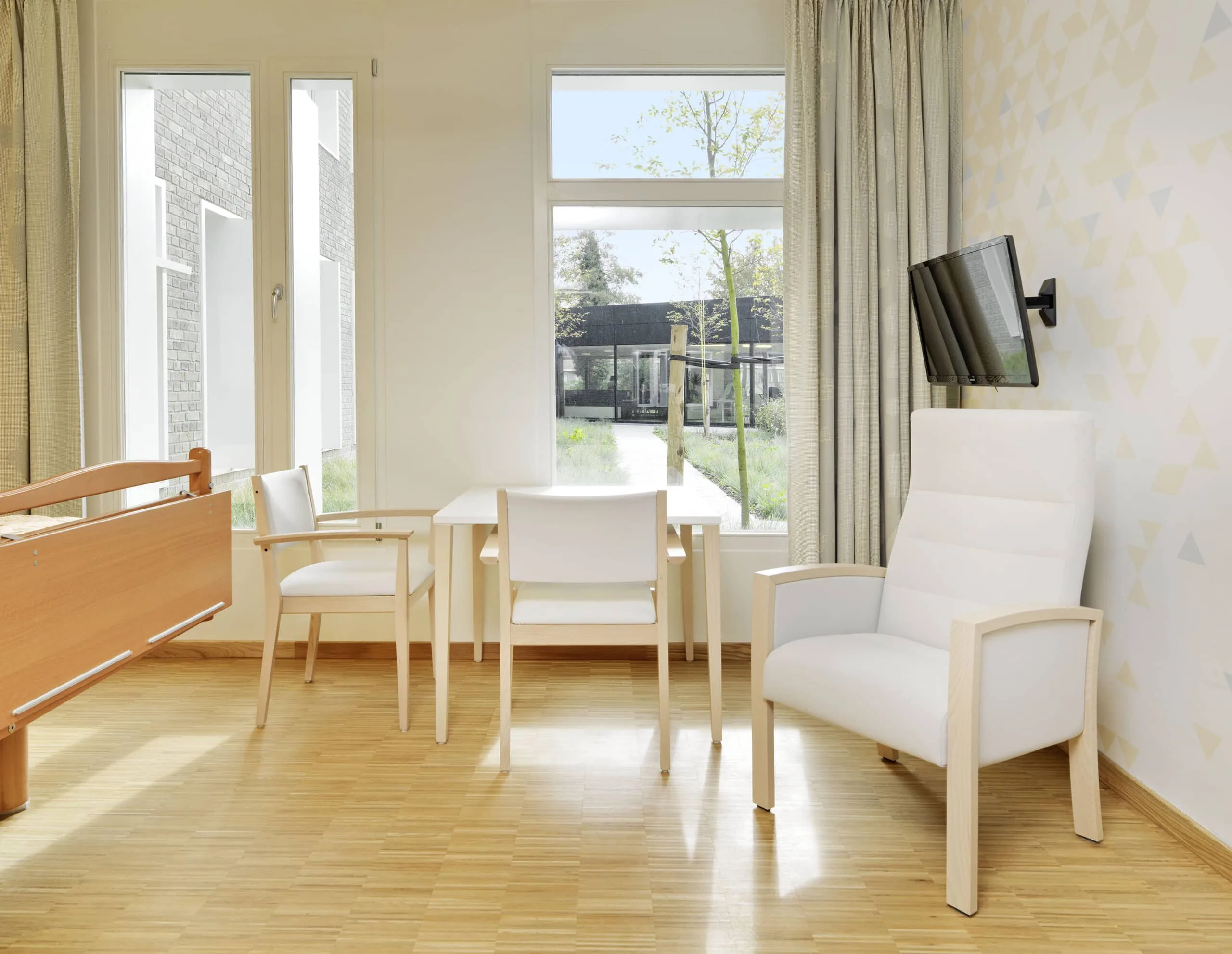 Helles Pflegezimmer mit großen Fenstern und Sitzgelegenheiten.