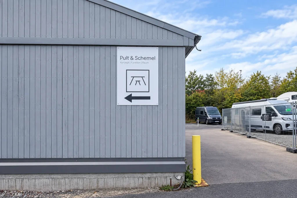 Schild von Pult & Schemel mit Pfeil nach links an der grauen Halle im Hof